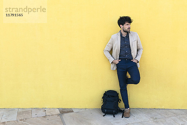 Junger Mann mit Rucksack vor gelber Wand stehend