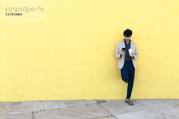Junger Mann mit Kaffee  um das Handy vor der gelben Wand zu überprüfen.