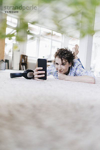 Frau liegt auf dem Boden in ihrer Wohnung  mit Smartphone