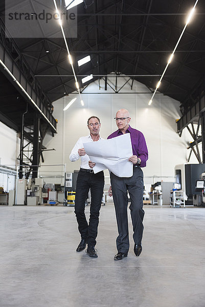 Zwei Geschäftsleute mit Plan zu Fuß in der Fabrikhalle