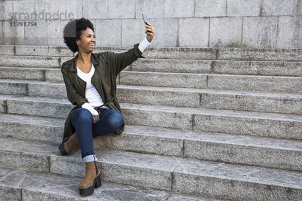 Lächelnde junge Frau sitzt auf einer Treppe und nimmt Selfie mit dem Smartphone.