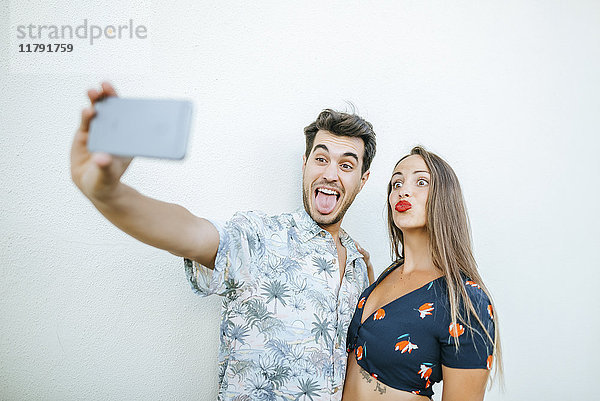 Verspieltes Paar  das einen Selfie mit Smartphone vor der weißen Wand nimmt.