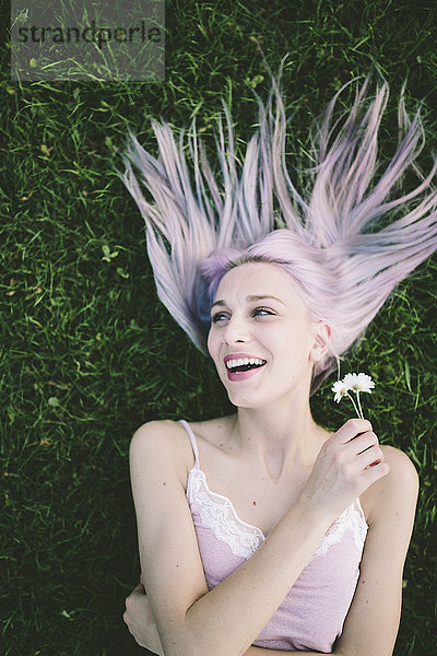 Porträt einer lachenden Frau  die mit geschlossenen Augen auf Gras liegt und Gänseblümchen hält.