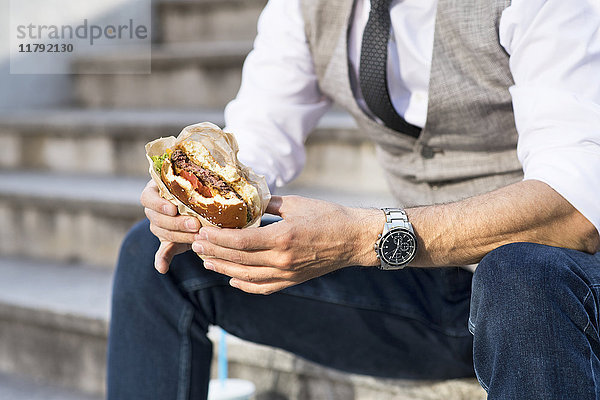 Geschäftsmann in der Stadt sitzt auf einer Treppe und isst einen Hamburger.