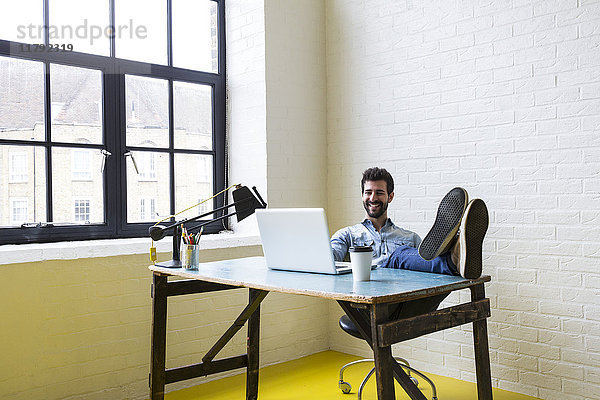 Lächelnder junger Mann sitzt mit erhobenen Füßen am Schreibtisch und schaut auf den Laptop.
