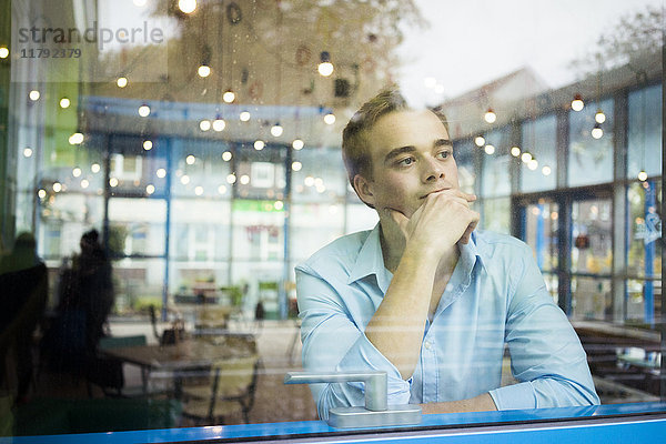 Porträt eines nachdenklichen jungen Mannes  der in einem Café sitzt und durchs Fenster schaut.