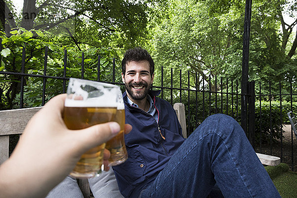 Porträt eines lächelnden jungen Mannes  der mit einem Glas Bier im Garten anstößt.