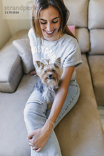 Lächelnde junge Frau sitzt mit ihrem Yorkshire Terrier auf der Couch.