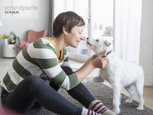 Lächelnde Frau sitzt auf dem Boden im Wohnzimmer und kuschelt mit ihrem Hund.