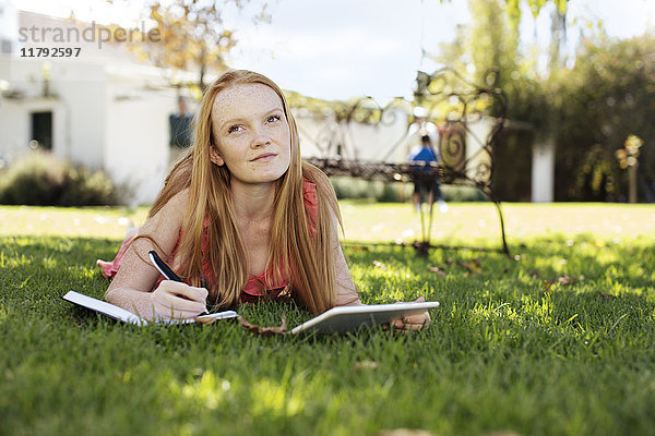 Grl mit langen roten Haaren im Gras liegend mit Tablette und Notizbuch
