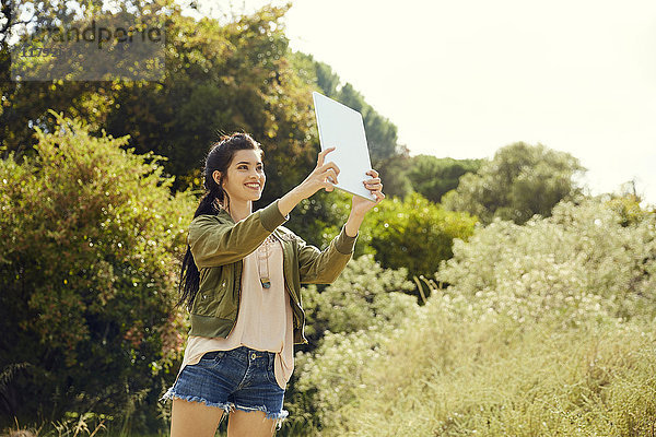 Lächelnde junge Frau nimmt Selfie mit Tablette in der Natur