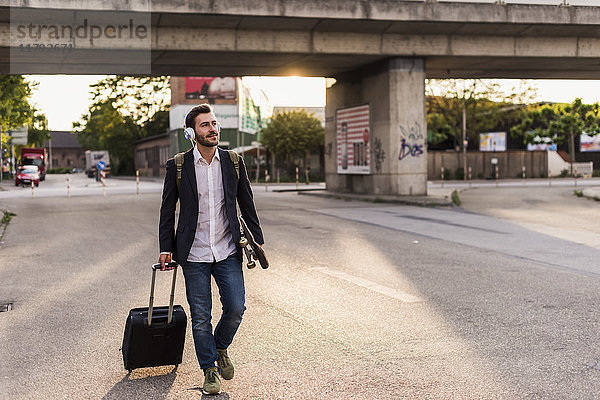 Junger Mann unterwegs mit Skateboard  Rollkoffer und Kopfhörer