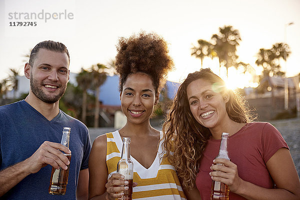 Gruppenbild von drei Freunden mit Bierflaschen am Strand bei Sonnenuntergang