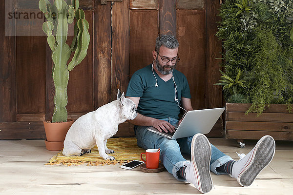 Der bärtige Mann sitzt mit seinem Hund zu Hause auf dem Boden und benutzt einen Laptop.