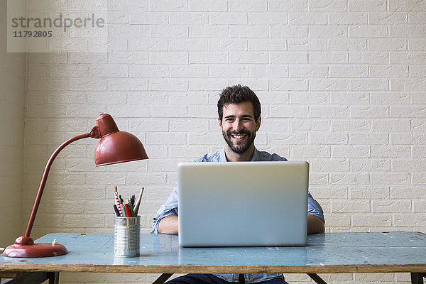 Porträt eines glücklichen jungen Mannes am Schreibtisch bei der Arbeit mit dem Laptop