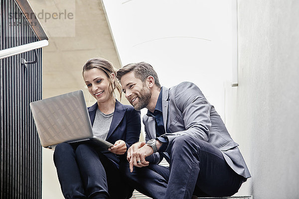 Geschäftsmann und Geschäftsfrau sitzen auf einer Treppe im Büro und teilen sich einen Laptop.
