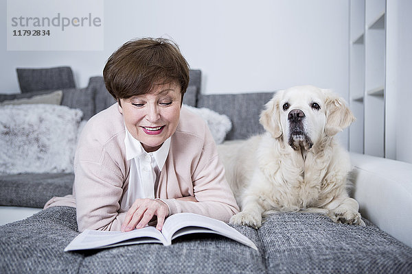 Seniorin auf der Couch liegend  Lesebuch mit Hund an der Seite