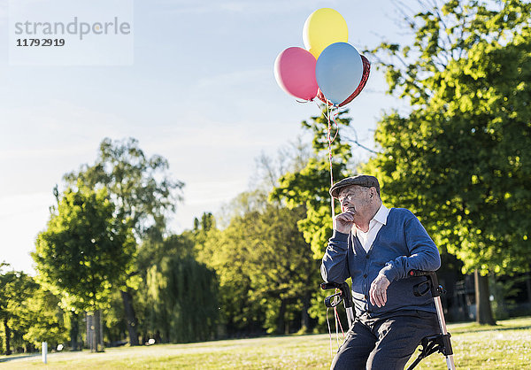 Nachdenklicher älterer Mann mit Luftballons auf seinem Rollator in der Natur.