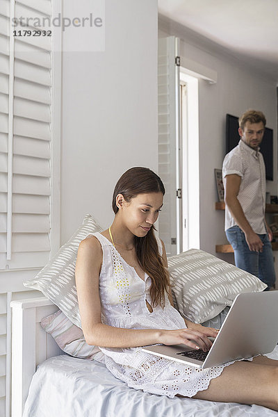 Junge Frau mit Laptop im Schlafzimmer mit Mann im Hintergrund