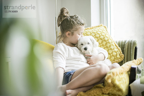 Kleines Mädchen sitzt auf einem Sessel zu Hause und kuschelt mit ihrem Hund.