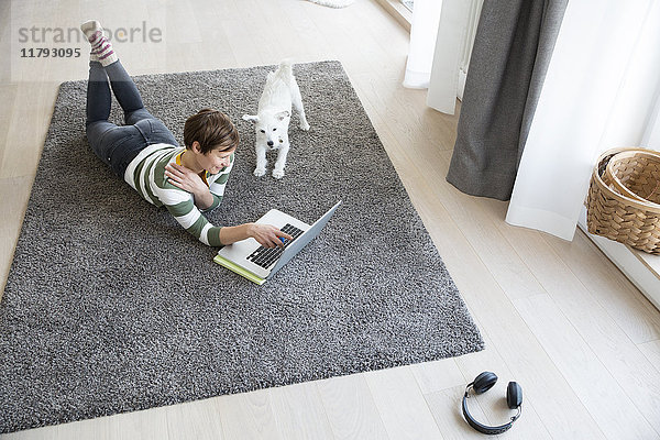Frau auf dem Boden liegend im Wohnzimmer mit Laptop  während der Hund sie beobachtet.
