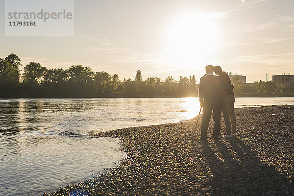 Rückansicht des älteren Mannes und seiner erwachsenen Tochter  die am Strand stehen und den Sonnenuntergang beobachten.