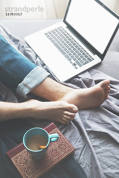 Mann auf Schlafcouch liegend mit Laptop und Tasse Kaffee  Teilansicht