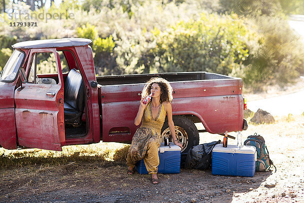 Junge Frau sitzt neben dem alten rostigen Pick-up und trinkt aus der Flasche.
