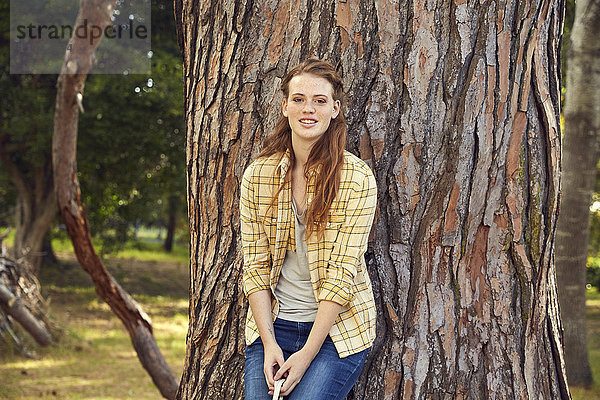 Porträt einer lächelnden jungen Frau  die sich an einen Baumstamm lehnt.
