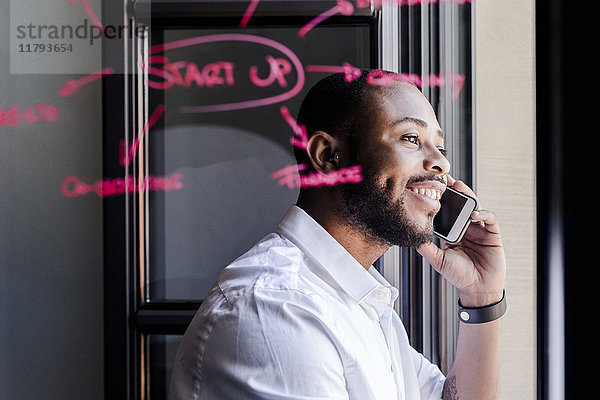 Lächelnder Geschäftsmann am Handy im Büro mit Schriftzug auf Fensterscheibe