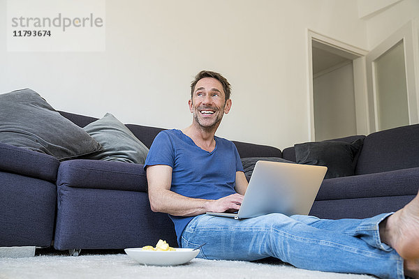 Porträt eines lachenden Mannes mit Laptop auf dem Boden im Wohnzimmer sitzend