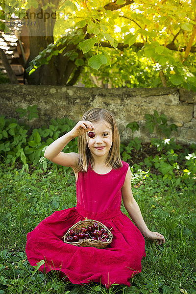 Porträt eines kleinen Mädchens in rotem Sommerkleid auf einer Wiese sitzend mit Kirschkorb