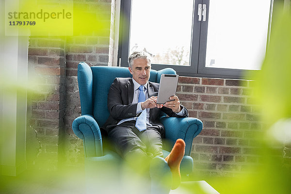 Der reife Geschäftsmann sitzt im Sessel und benutzt ein digitales Tablett.