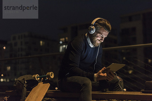 Lächelnder junger Mann mit Tablette und Kopfhörer in der Stadt bei Nacht