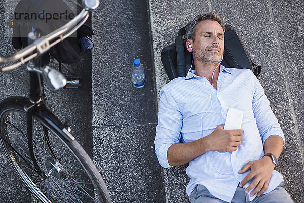 Mann mit geschlossenen Augen auf Treppe liegend mit Handy und Ohrstöpseln neben Fahrrad