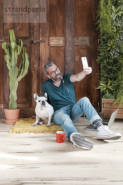 Bärtiger Mann sitzt mit seinem Hund auf dem Boden zu Hause und nimmt Selfie mit dem Smartphone.