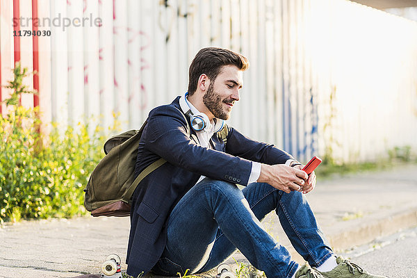 Lächelnder junger Mann sitzt auf dem Bürgersteig und überprüft sein Handy.