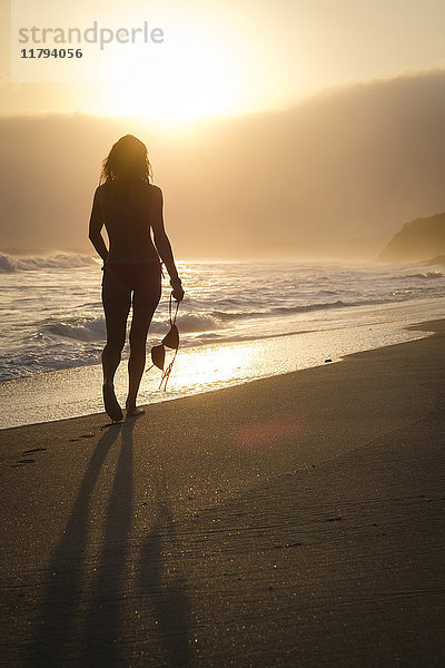 Mexiko  Riviera Nayarit  Silhouette einer Frau  die an einem Strand in den Sonnenuntergang geht  während sie ihr Bikini-Top hält.