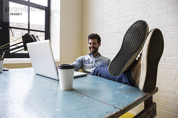 Lächelnder Mann sitzt mit erhobenen Füßen am Schreibtisch und schaut auf den Laptop.