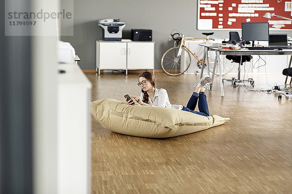Junge Frau mit Handy in Bohnentüte im Büro liegend