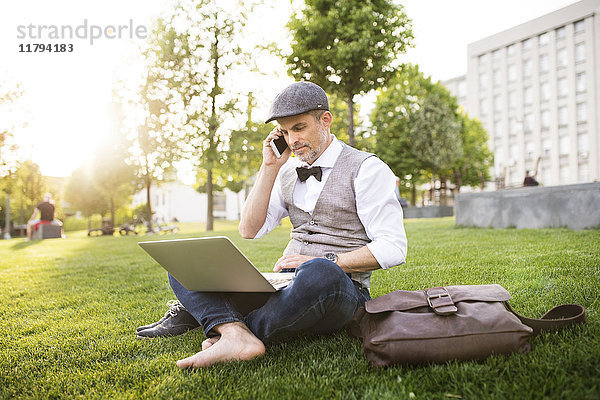 Selbstbewusster Geschäftsmann mit Laptop und Smartphone im Stadtpark auf Rasen sitzend