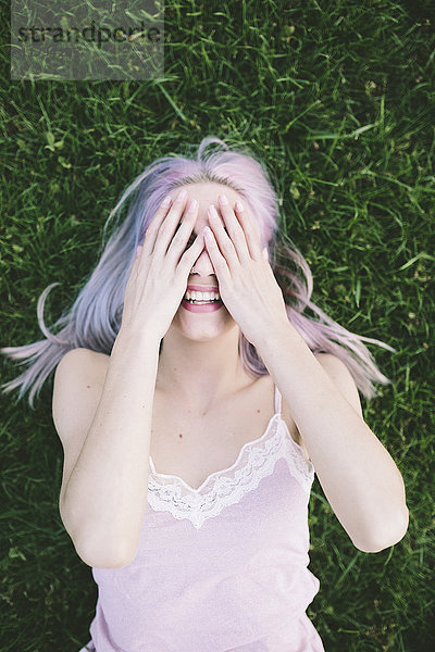 Lachende Frau liegt auf Gras und bedeckt die Augen mit ihren Händen.
