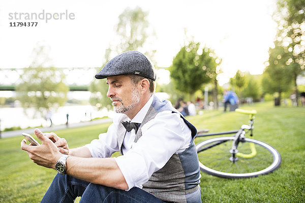 Selbstbewusster Geschäftsmann mit Fahrrad und Smartphone im Stadtpark auf Rasen sitzend