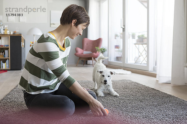 Lächelnde Frau  die zu Hause auf dem Boden sitzt und mit ihrem Hund spielt.