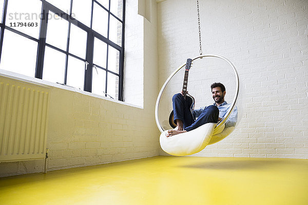 Lächelnder Mann mit Gitarre sitzt auf dem Swing in seinem Loft