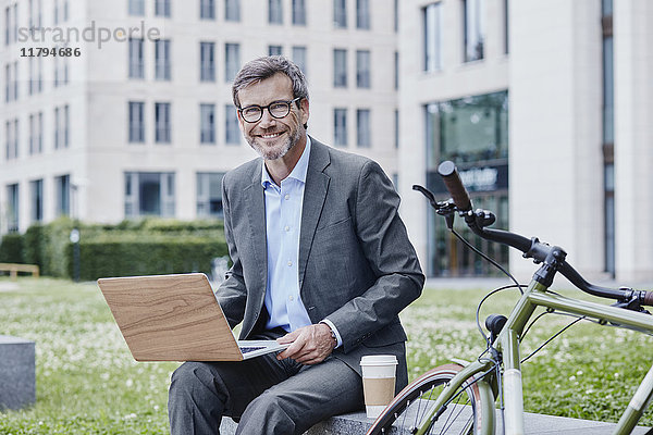 Porträt eines lächelnden Geschäftsmannes im Freien mit Laptop  Kaffee zum Mitnehmen und Fahrrad