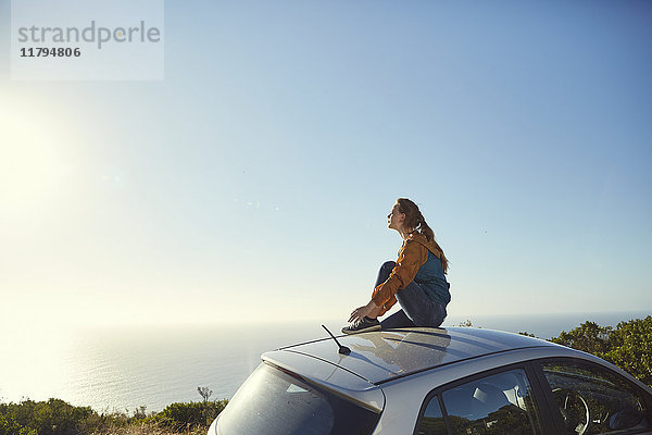 Südafrika  Kapstadt  Signal Hill  junge Frau sitzt auf dem Auto und genießt den Blick aufs Meer.