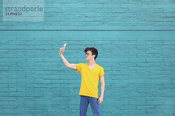 Junger Mann nimmt einen Selfie mit Smartphone vor der blauen Ziegelwand