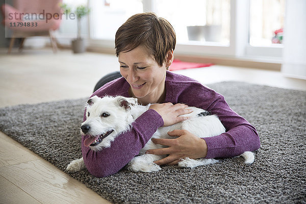 Frau  die auf einem Teppich im Wohnzimmer liegt und ihren Hund umarmt.