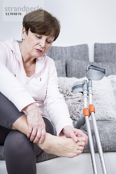 Seniorenfrau mit Krücken auf der Couch  die ihren Knöchel überprüft.